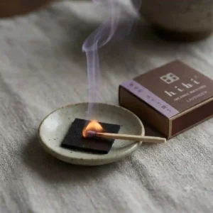 An illustration of a Hibi incense match resting horizontally on its specialized round, black heatproof mat while gently smoking.
